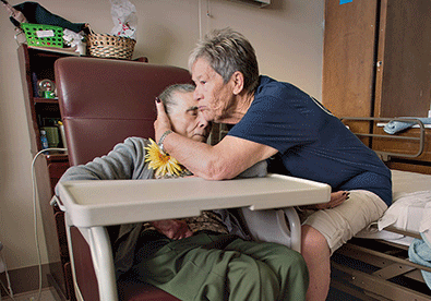 a senior woman sits in a chair with a tray while another woman hugs her head and consoles her after understanding the definition of hospice care