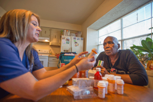 a nurse sits with a senior woman at a table with medicine bottles and the nurse is answering the woman's home health care faqs
